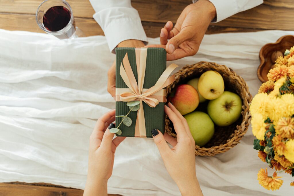 Hands exchanging a wrapped gift over a Thanksgiving table setting with apples and flowers.